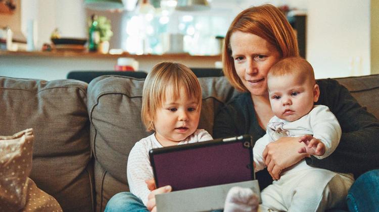 Mother sitting with toddler and infant on a couch. They are interacting with a tablet