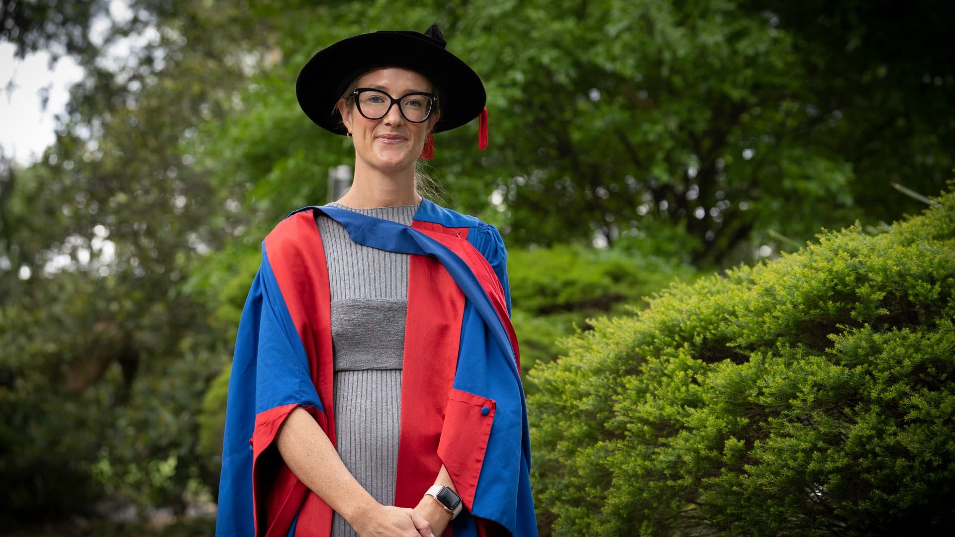 Dr Carmen Naylor, wearing a grey dress and the blue and red graduation gown, stands in front of a green, bush background. Photo: Paul Jones
