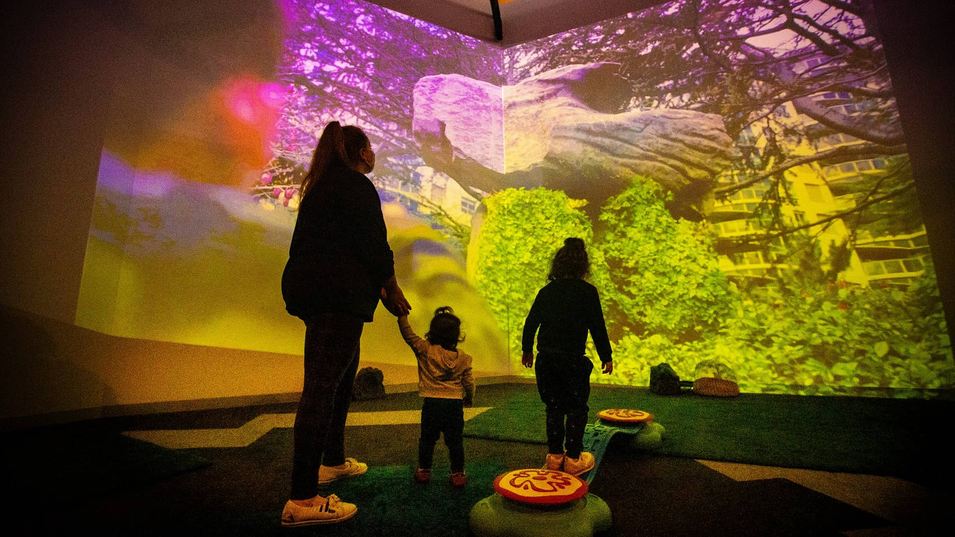 A mother holds the hand of her child while another child watches a large screen. They are silhouetted against the vibrant colours of the screen. Photo: Paul Jones