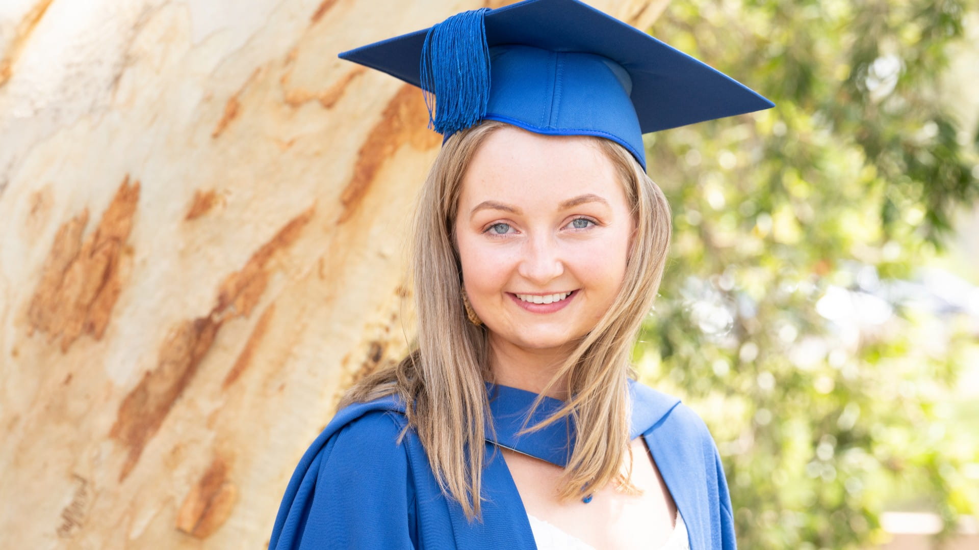 Emily Nield wears a blue cap and gown and stands in front of a tree. She is smiling. Photo: Paul Jones