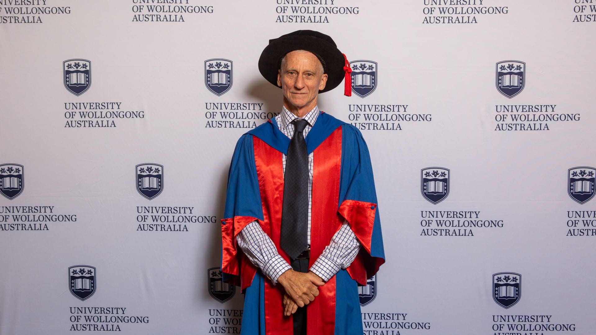 Professor Paul Else wears a red graduation gown and stands in front of a UOW wall. Photo: Andy Zakeli