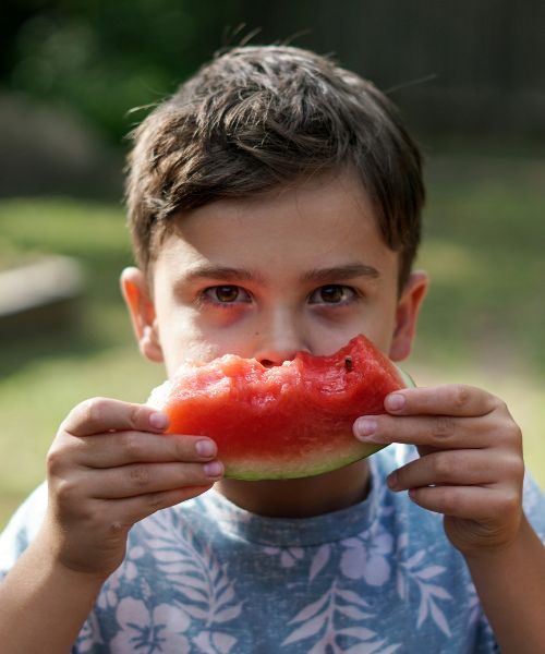 A young boy holds a piece of watermelon in front of his face