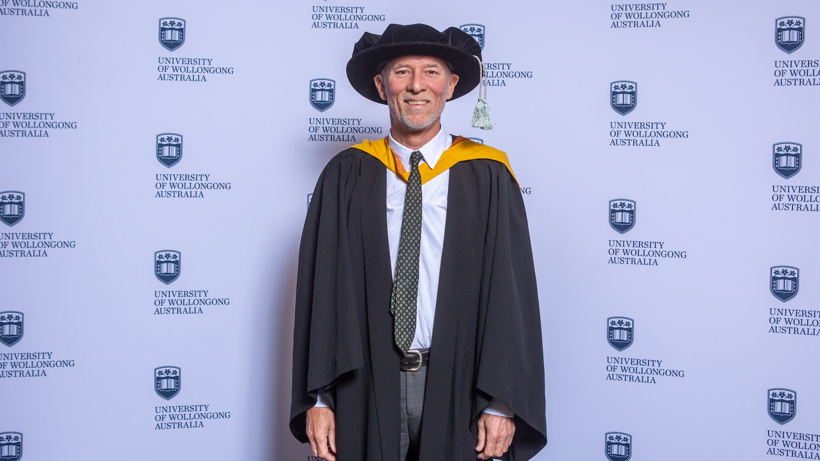 Professor Frank Deane in a black and gold gown and cap in front of a UOW media wall. Photo: Andy Zakeli