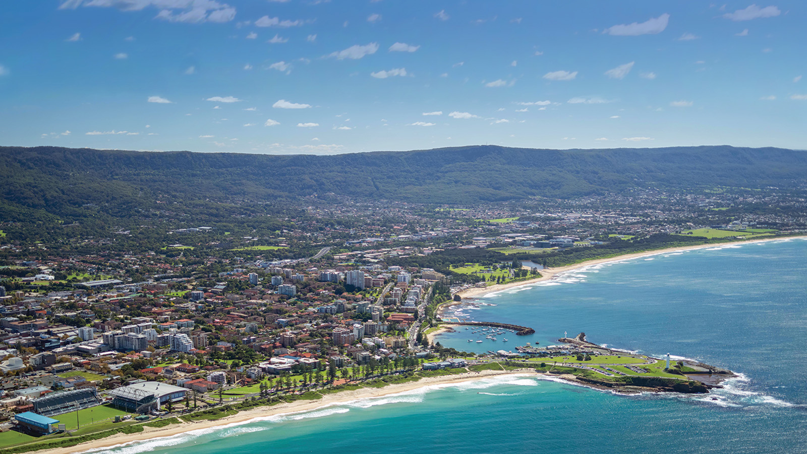 Aerial view of Wollongong city with a beach, lighthouse, and green hills in the background.