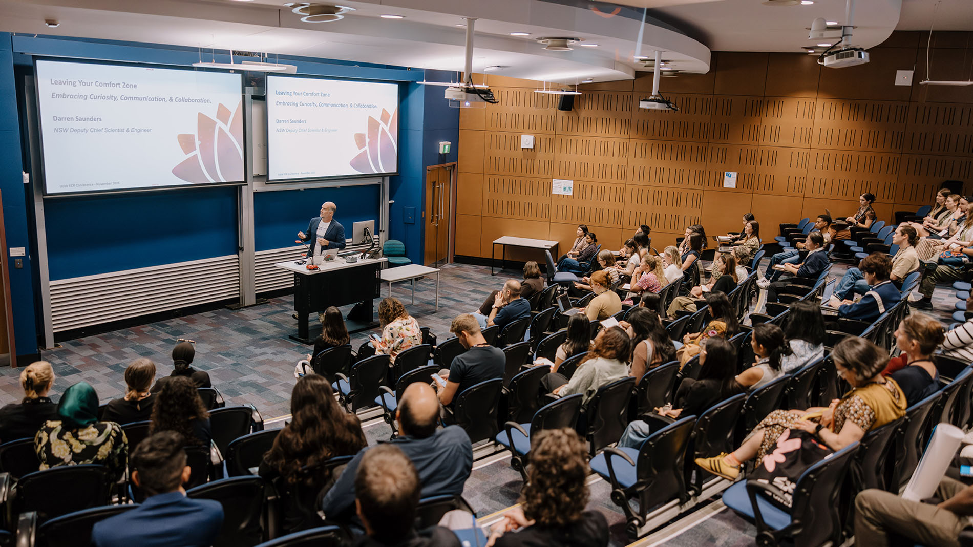 A lecturer speaks to a seated audience in a modern classroom, with presentation slides displayed on two large screens at the front.