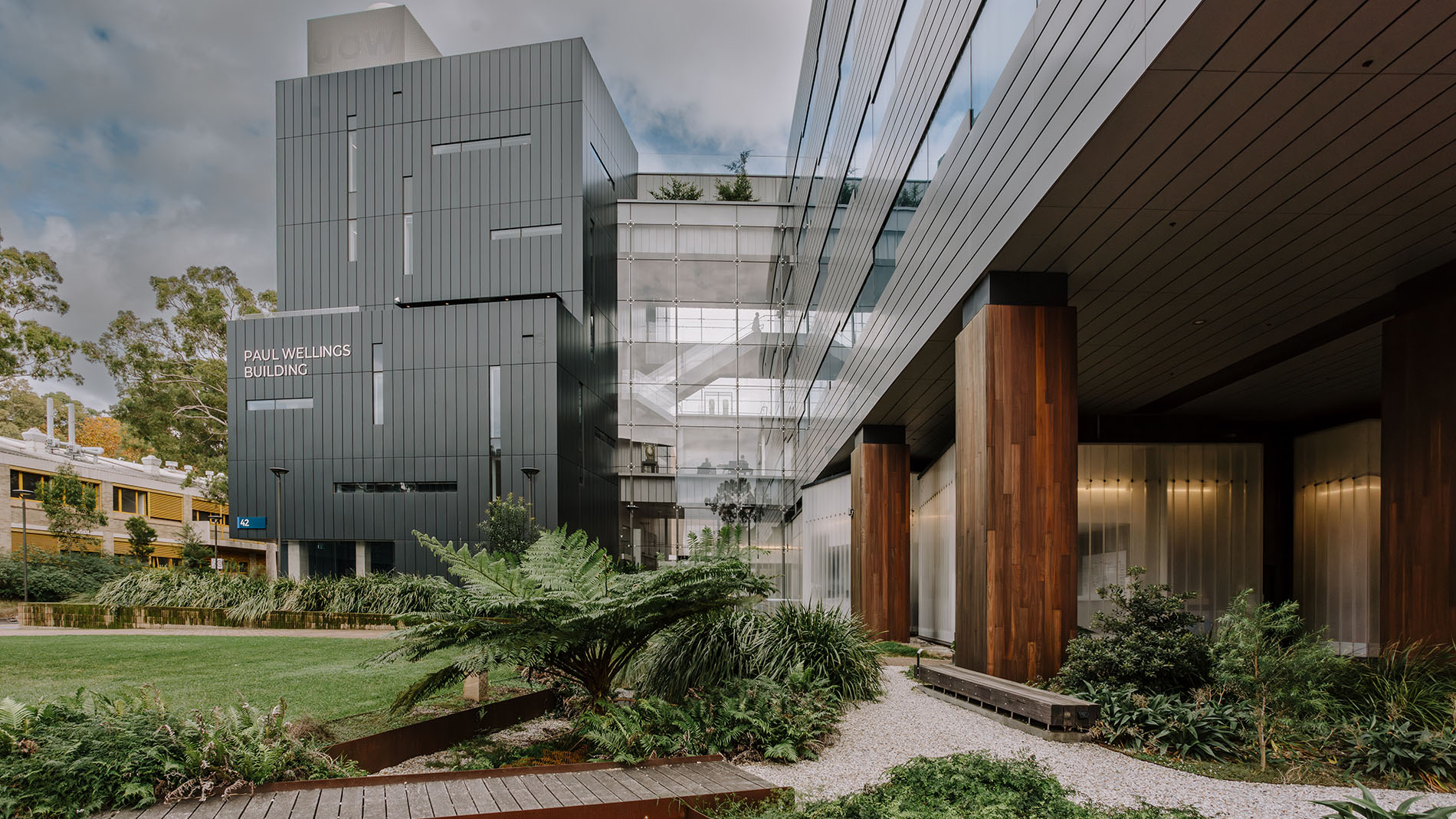 Exterior view of the University of Wollongong’s Paul Wellings Building, a contemporary multi-storey structure with dark metal cladding, large glass façades and timber architectural features. Landscaped gardens, native plants and a lawn surround the building, with the “Paul Wellings Building” sign visible on the exterior wall.