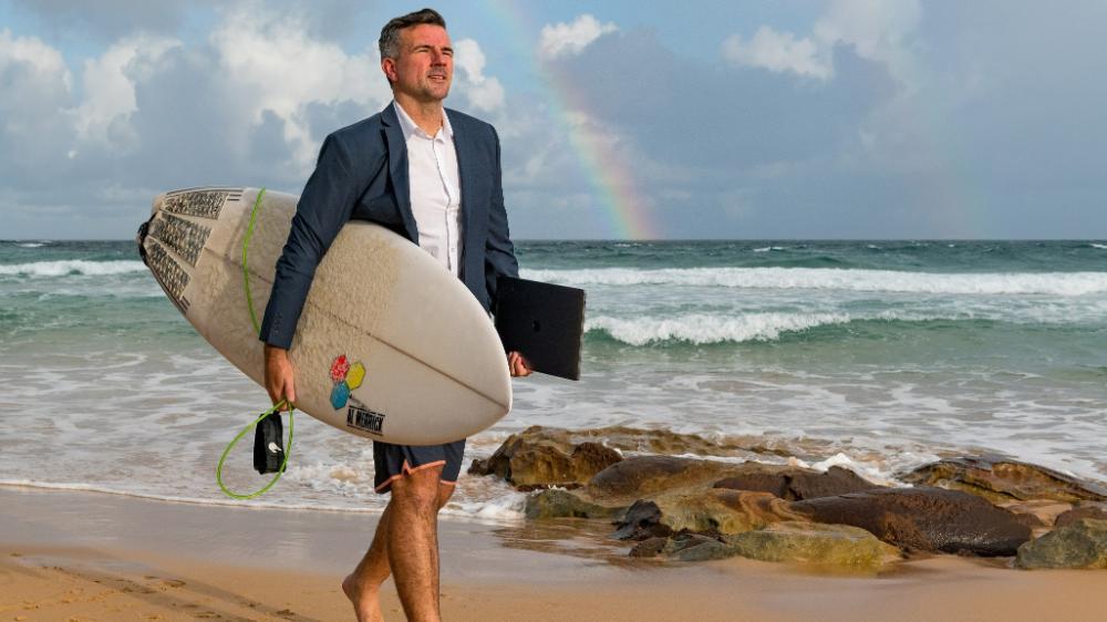UOW graduate Dane Sharp with surfboard and laptop walking on beach