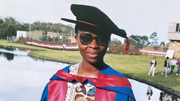 Omowunmi Sadik in front of teh duck pond at her 1994 graduation. She wears a black grad hat and a blue and red cloak
