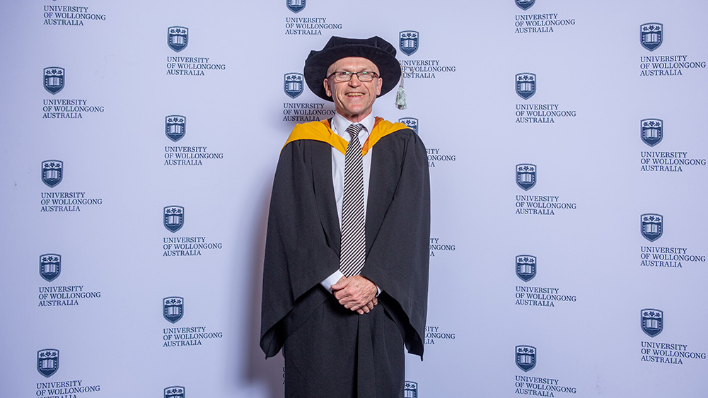 Professor Paul Cooper wears a black graduation cap and gown and stands in front of a UOW wall.