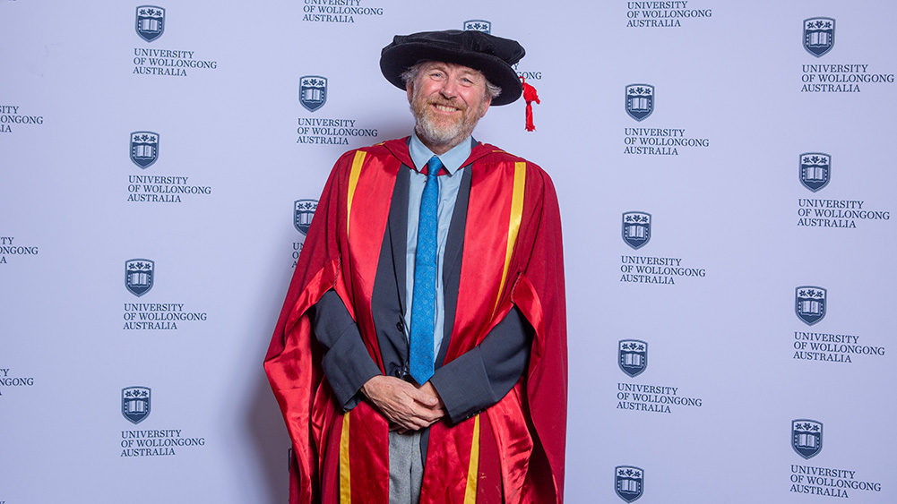 Professor Will Price wears a black graduation cap and red gown and stands in front of a UOW wall.