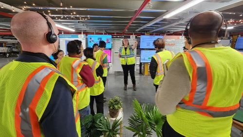 A group of people standing in front of a screens and an instructor. All are wearing yellow safety vests and headphones.