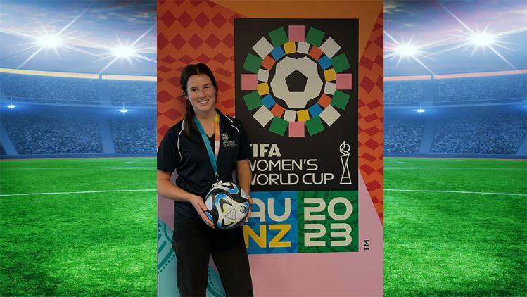 Emily Wescombe, Marketing student/intern standing in front of a FIFA Womens World Cup backdrop. She is also holding a soccer ball.