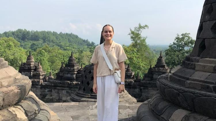 Maya Willis wearing a yellow top and white pants while smiling standing in front views of Indonesia.