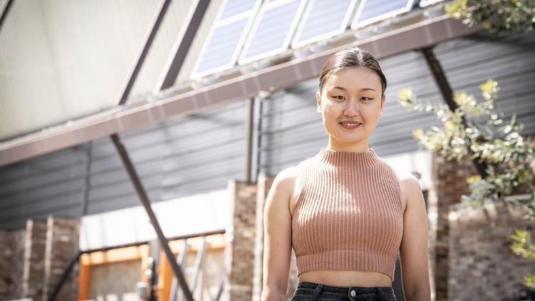 Student smiling, wearing a brown top and standing outside the SBRC building with solar panels.