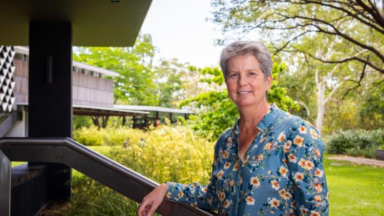 Sarah Larkins Keynote Speaker Rural Health Conference 2023, wearing a floral nd blue top, leaning against a railing under an outside cover. Green trees are in the background.