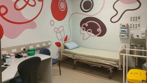 Medical consultation room with a bed and a desk with two chair. Colourful patterns appear on the wall.