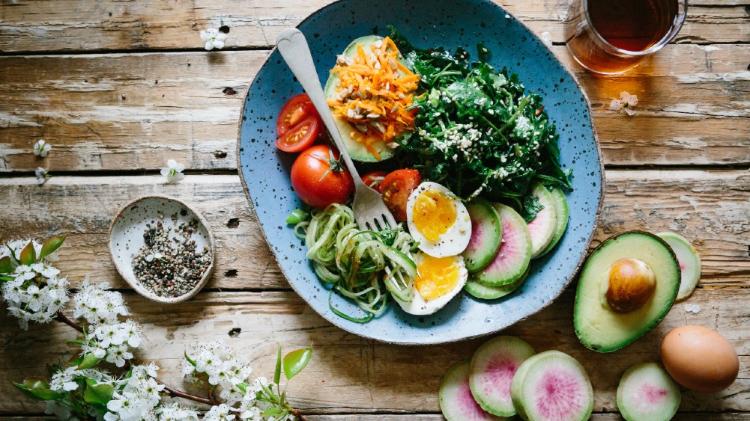 Plate with nutritious food including eggs and tomatoes with a fork sitting in the plate. A cup of tea and a half  of an avocado are also on the wooden boards and a white flower.