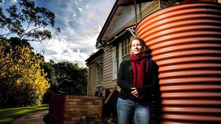 Carrie Wilkinson stands against a orange water tank, with her hands infront. Carrie wear a navy sweater, blue denim jeans and a maroon scarf