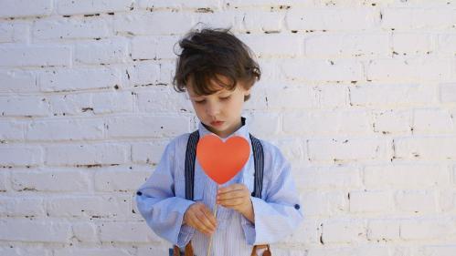 Young boy standing infront of brick wall, holding a red cardboard heart