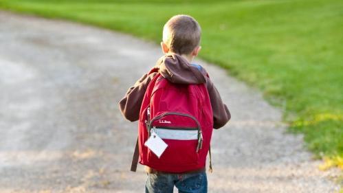 Boy wearing brown jumper, blue jeans and a red back pack walking away into distance