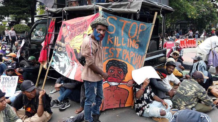 West Papuan man stands on the street, holding a canvas sign against a truck. The Sign says 