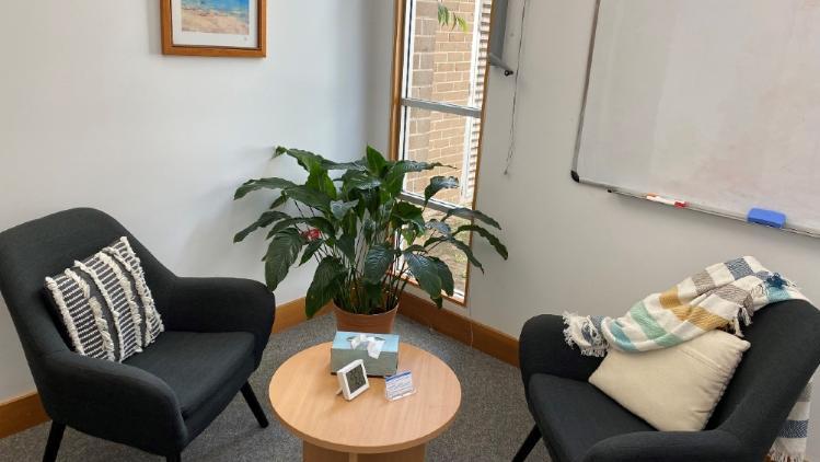 A therapy space with two black sitting chairs with pillows around a low rounded table with a grey tissue box on it. A seaside picture and whiteboard hangs on the walls and a plant in a pot in front of a window.