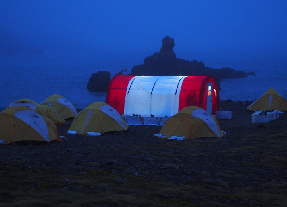 A number of tents pitched on rocky ground, with the ocean in the background.