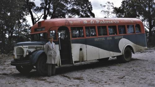 Man in beige work coat standing near red-topped Rutty's bus at Summit Park, Mt Keira.