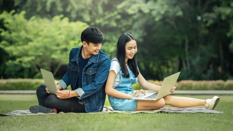 Two people sitting on grass with laptops smiling.