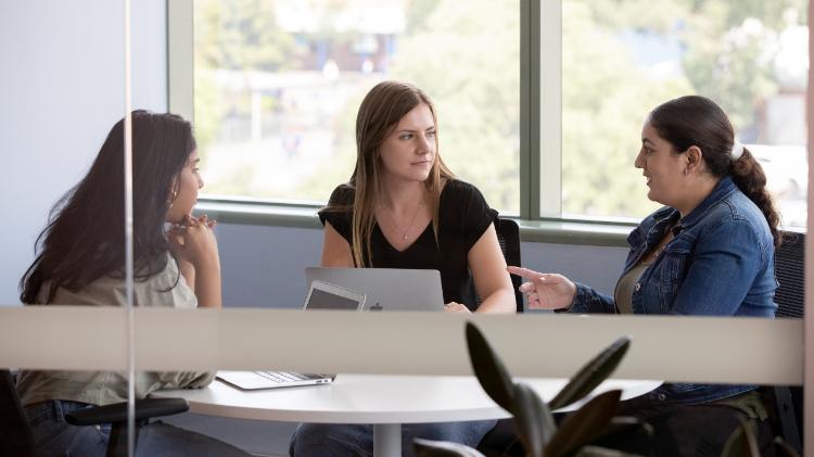 Three students sitting together at a table talking with a window behind them.