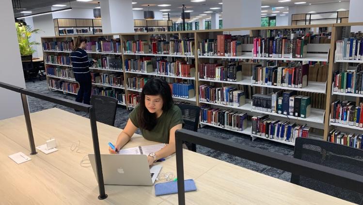 Student studying at table in library level one. Student browsing book shelves.
