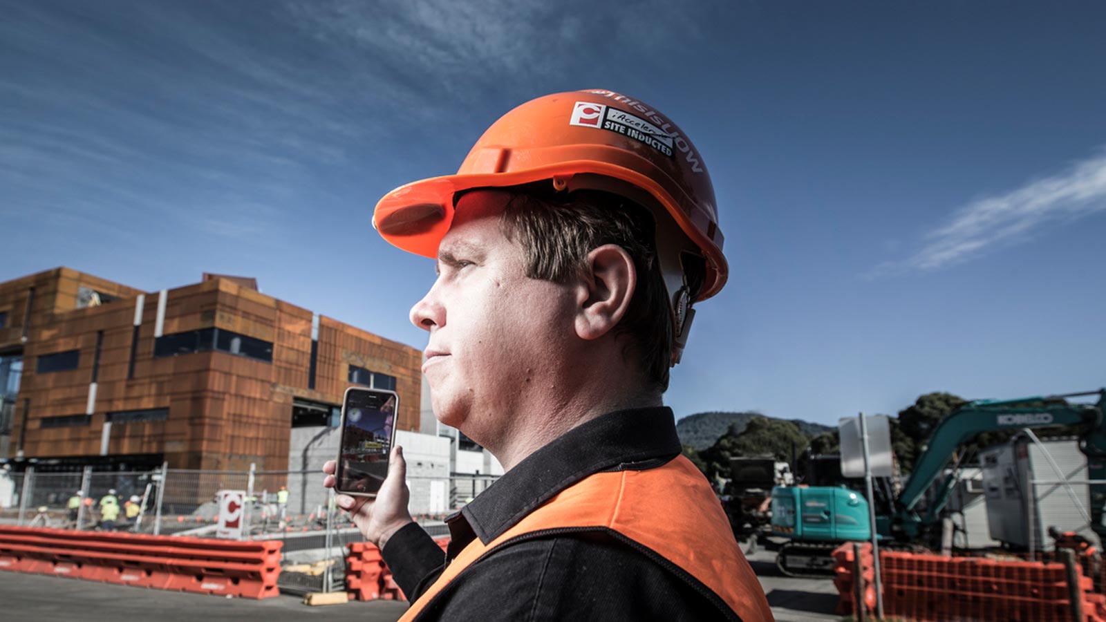 A man in an orange hard hat and vest, holding a smartphone towards a costruction site.