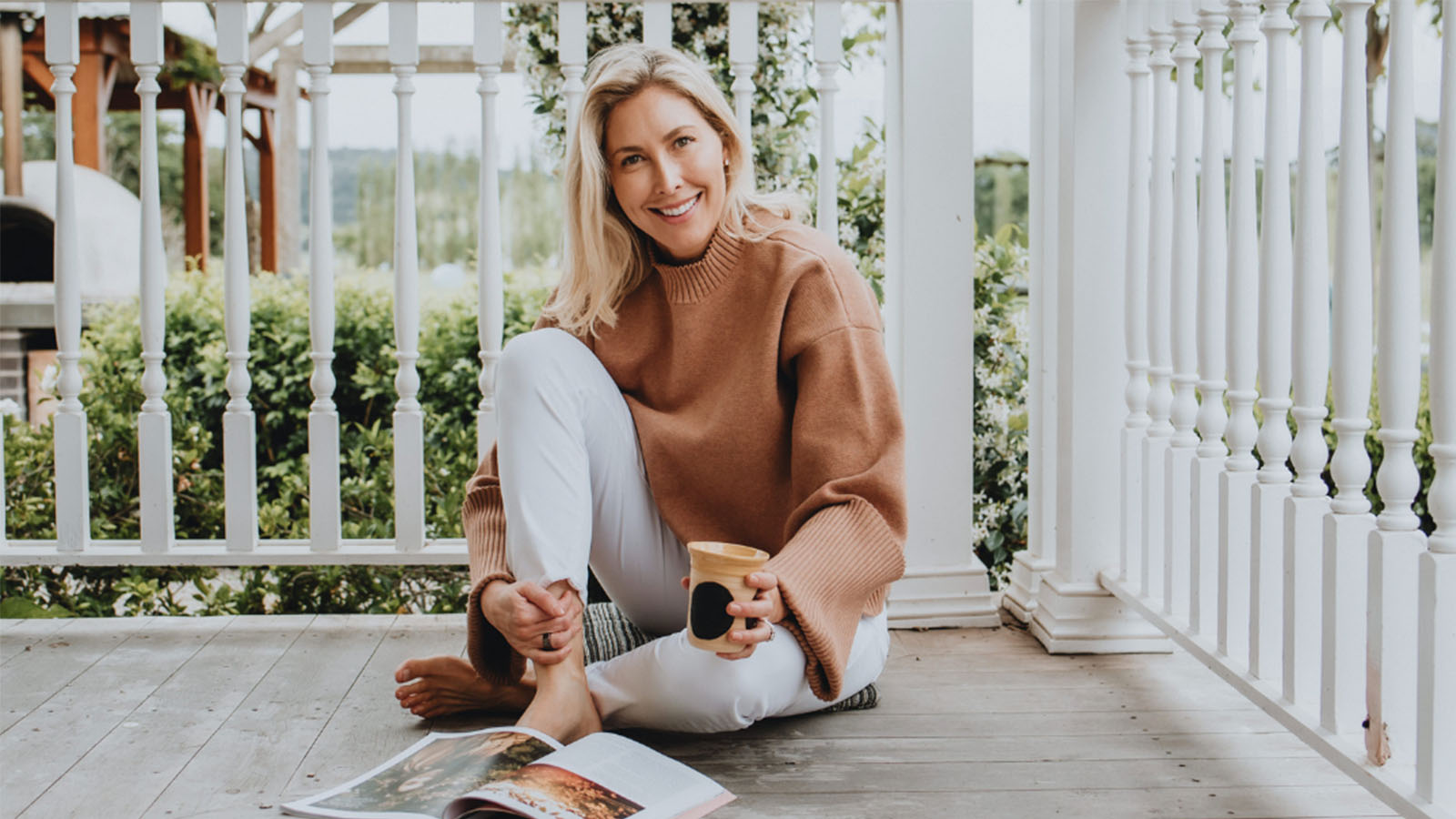 A woman with blonde hair is wearing a brown jumper and white pants. She is sitting on the floor of a verandah, smiling.