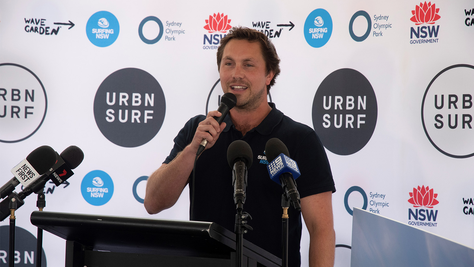 Luke Madden is speaking at a press conference. He is wearing a black t-shirt, holding a microphone in front of a Surfing NSW media wall. There is a black podium in front of him with microphones placed on it.