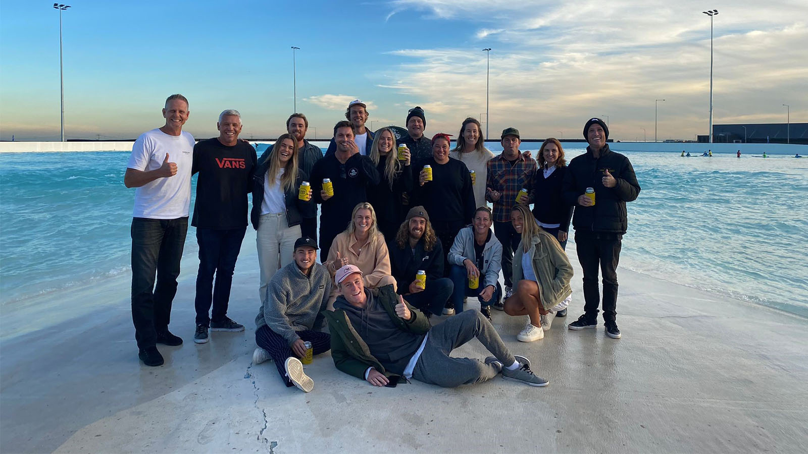 A group of people are standing on a bank in front of blue water, holding yellow cans.