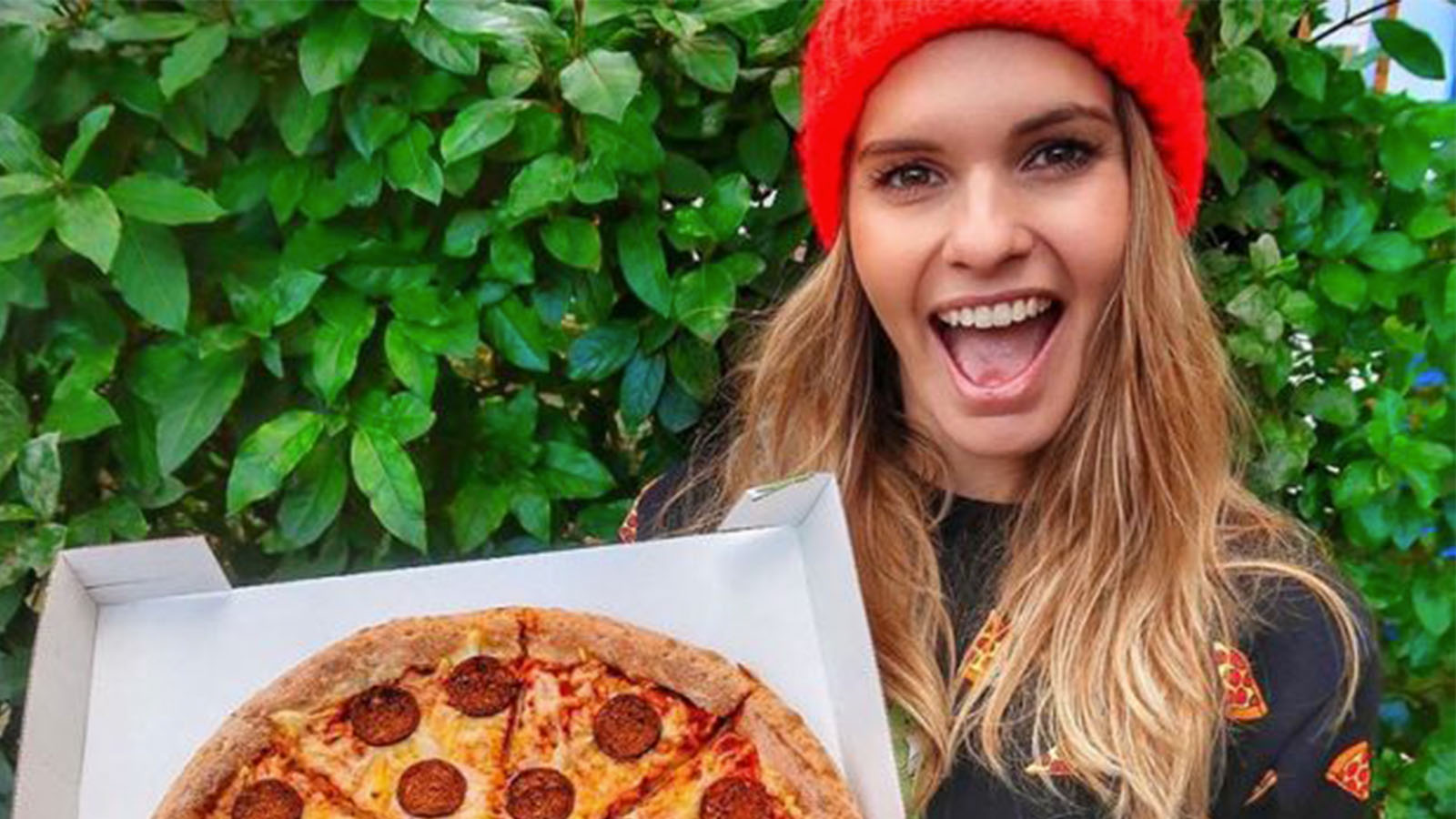A woman with light brown hair is wearing a red beanie. She is holding up a pizza and smiling.