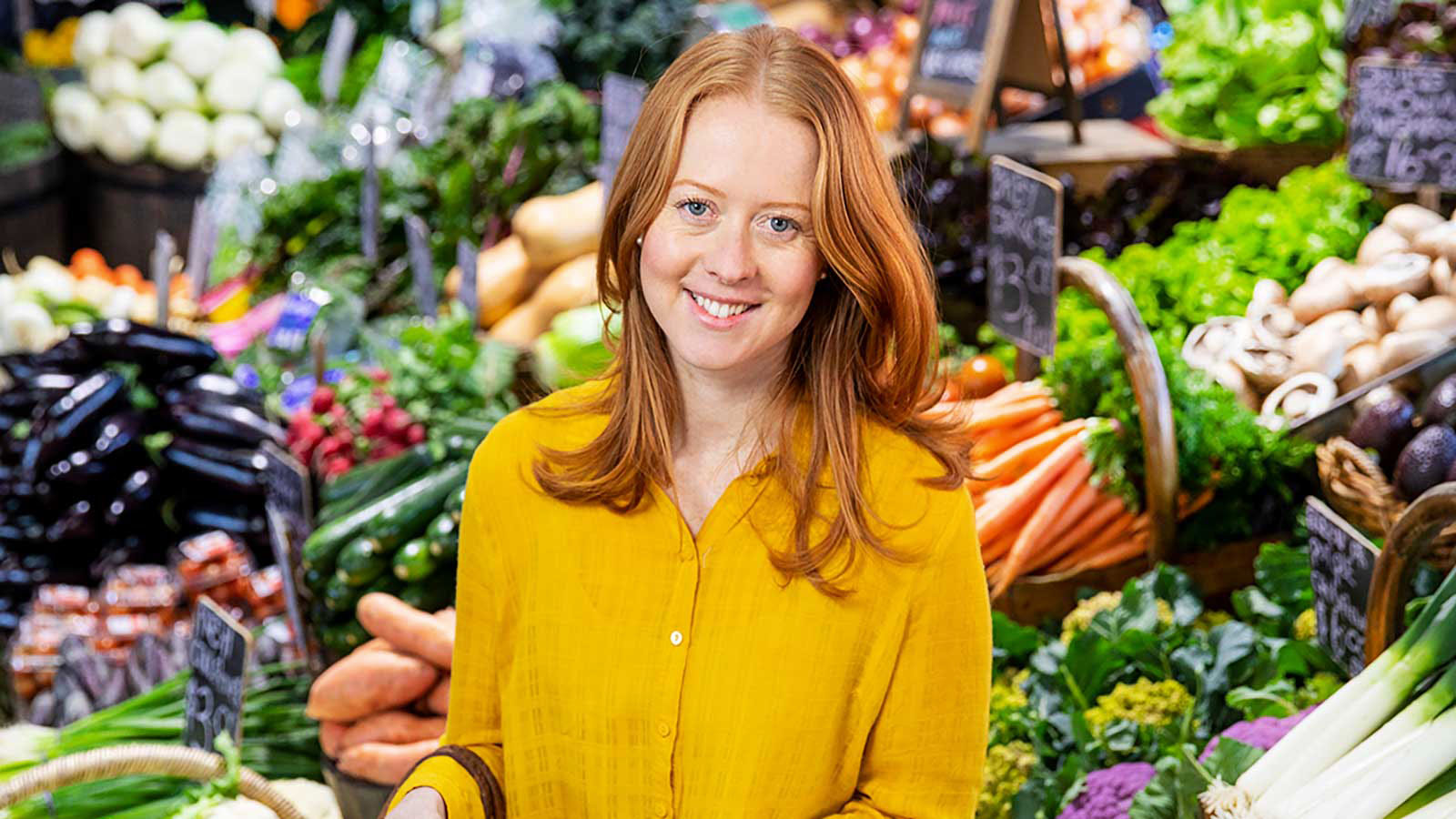 A woman with red hair and a yellow top is standing in front of a fresh produce display