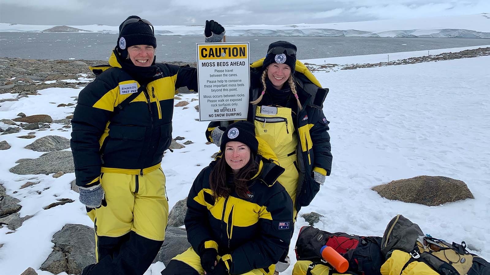 Three women in thermal black and yellow overalls are standing in front of a caution sign in Antarctica