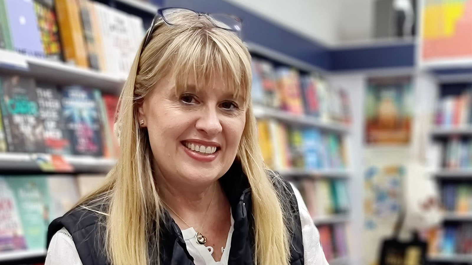 A woman with blonde hair is standing in a bookshop, smiling.