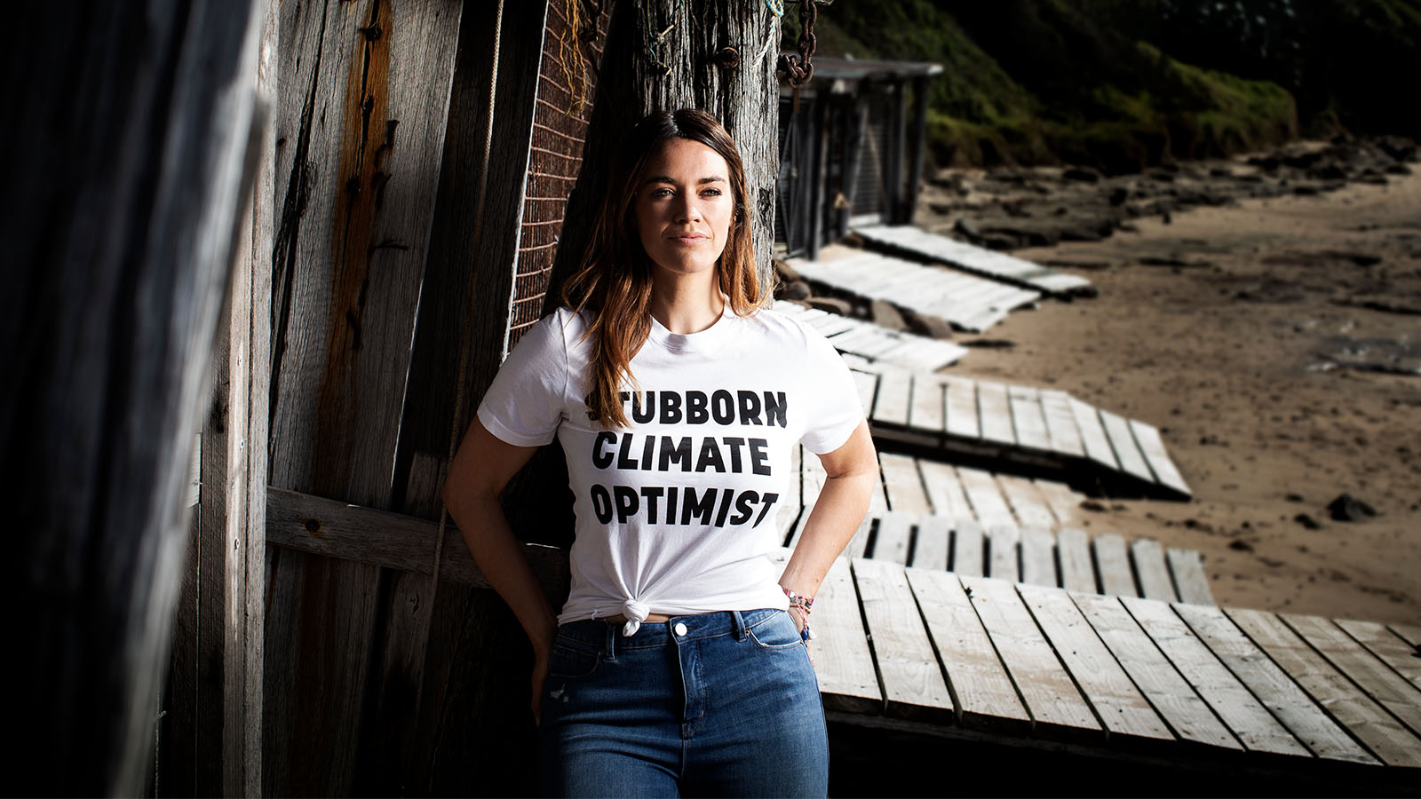 A woman is standing on a beach. she is wearing blue jeans and a white shirt that reads 'Stubborn climate optimist'