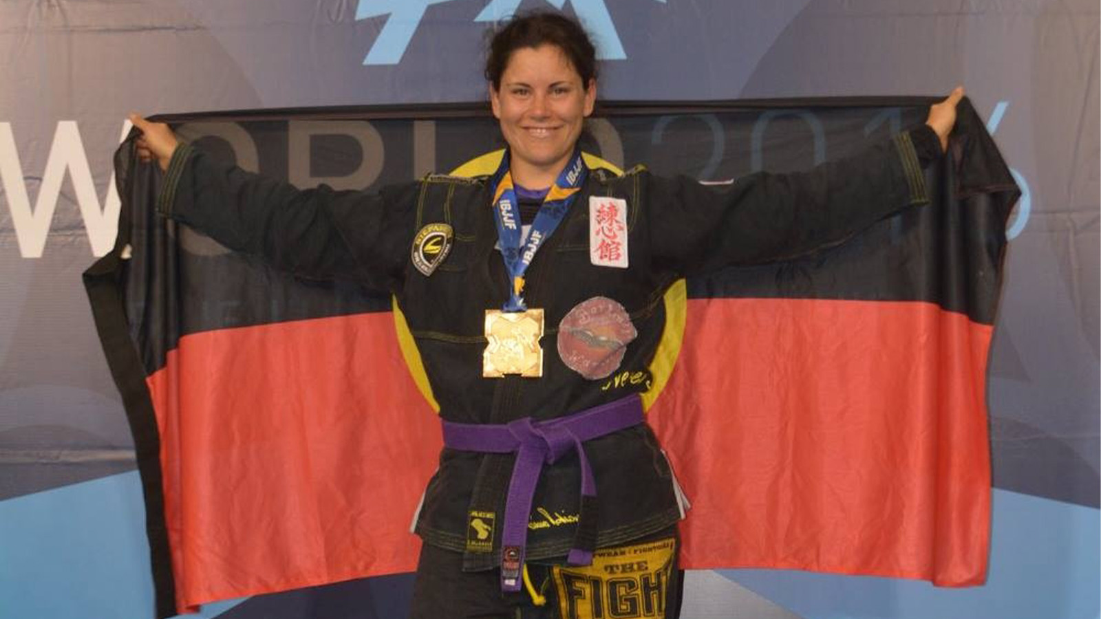 Shantelle Thompson is wearing a black jiu-jitsu uniform with a purple belt. She standing in front of a World Championships 2016 sign, smiling, holding an Aboriginal flag up behind her back.