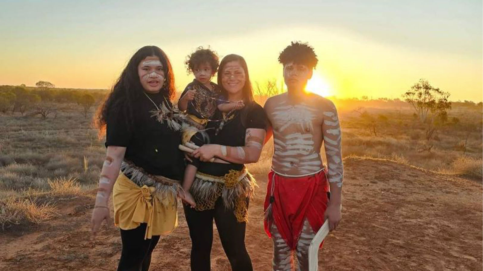 Shantelle Thomspon is standing against a sunset, holding a small child, with two teenagers by her side. The family is all wearing traditional Aboriginal body and face paint, and weaved skirts.