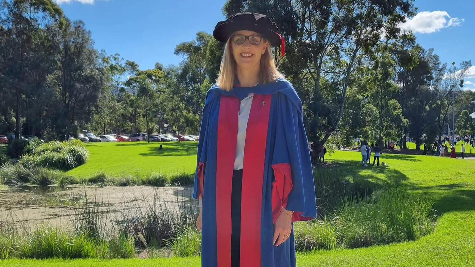 A woman is standing in a blue and red graduation gown and black doctorate graduate hat on the UOW lawn.