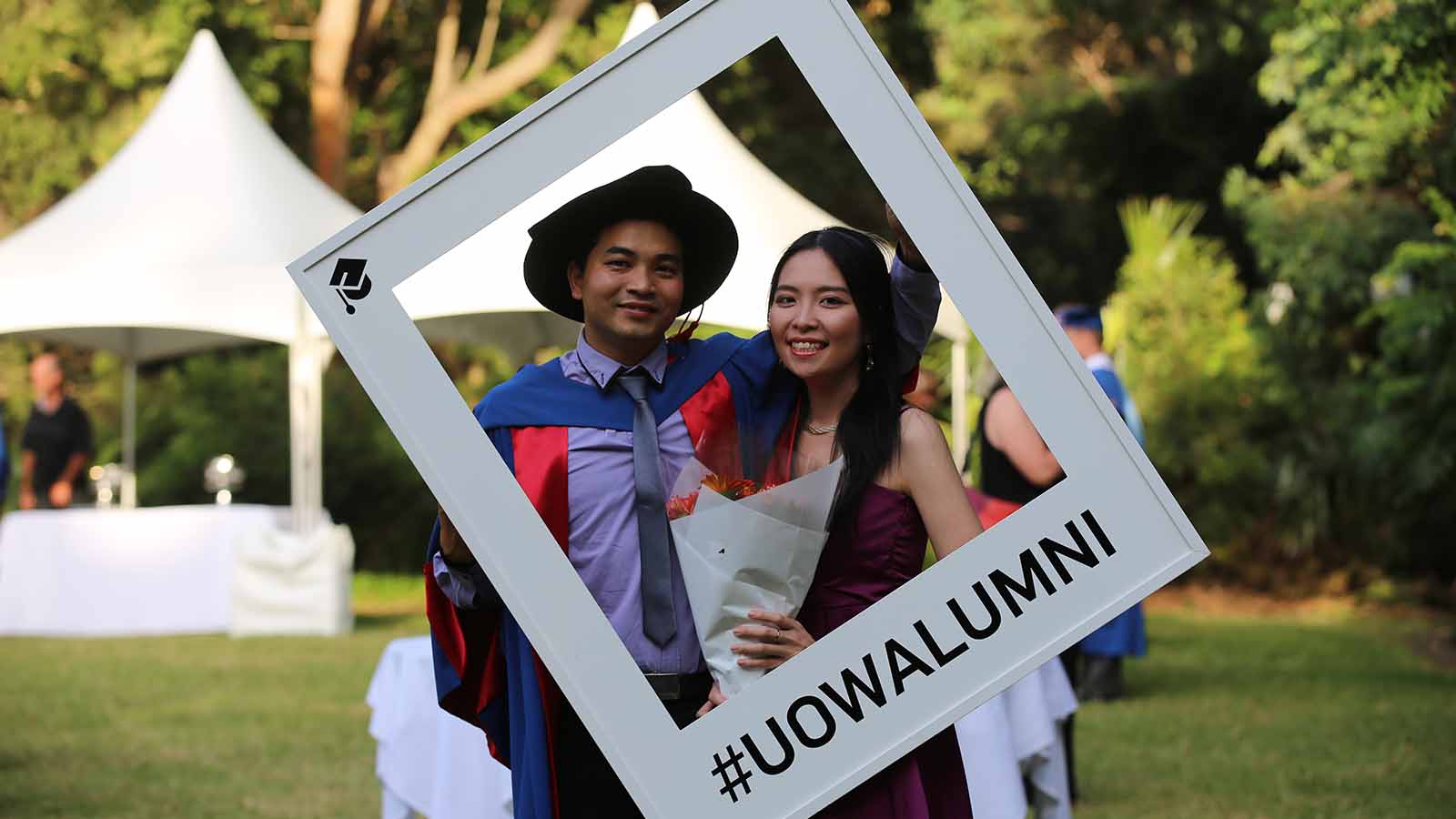 Dr Thong Pham is at his graduation with a woman, holding a corflute grame with #UOWALUMNI written on it