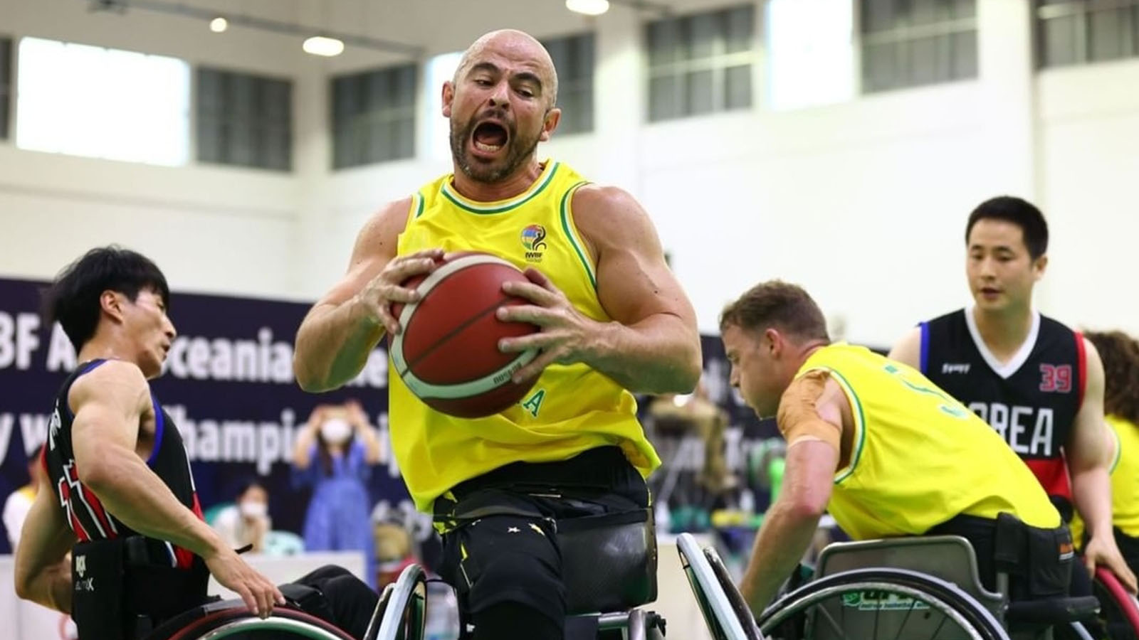 A man is playing wheelchair basketball. He is wearing a yellow and green basketball singlet, holding a basketball in both hands.