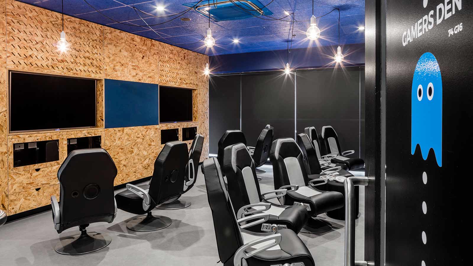 Inside of a UOW Accommodation game den. There is an exposed brick wall, black wall and blue ceiling. There is an array of gaming chairs across the grey floor.