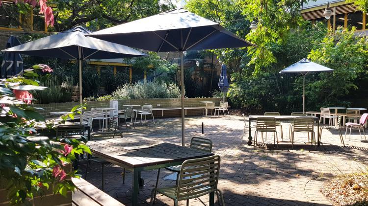 Chairs and tables shaded by trees and umbrellas in a brick courtyard.