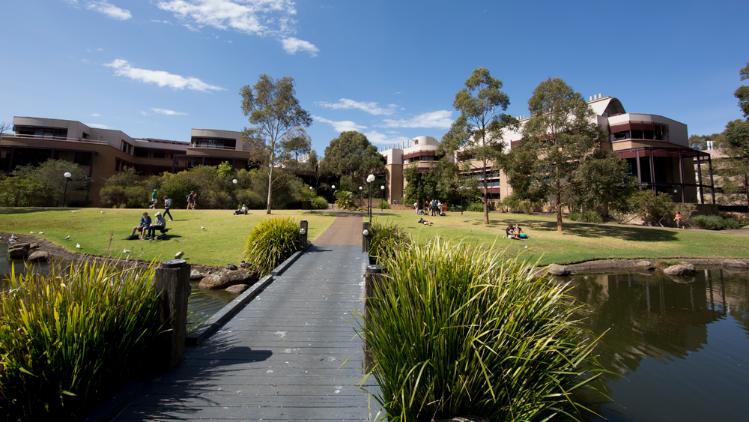 A pathway leads across a stream of water towards green grass and buildings on campus at UOW.