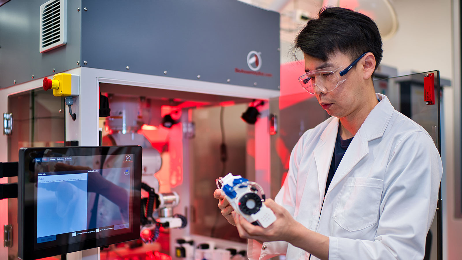 A man in is standing in a lab wearing a lab coat and safety goggles. He is holding a white device connected to a 3D printer.