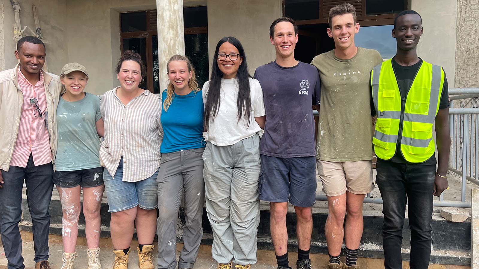 A group of people standing in front of a beige building. They have paint and plaster on them from renovating.